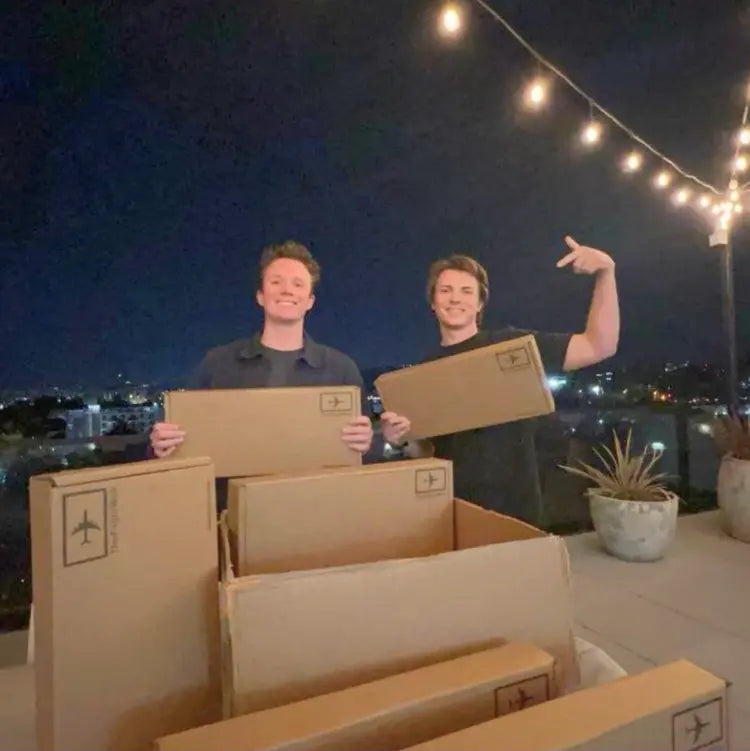 FlightWall founders Aaren and Alex holding FlightWall boxes on a rooftop at night with string lights and cityscape in the background.